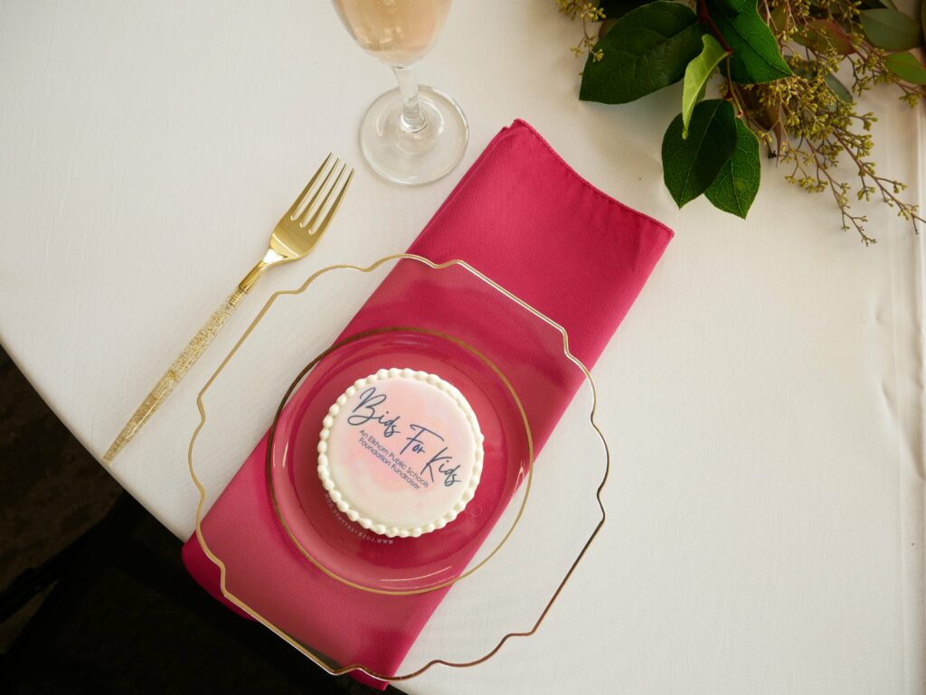 A table setting featuring a golf fork, glass of wine, clear plate, pink napkin, and a bit of greenery off camera. On the plate sits a cookie with the Bids For Kids logo.